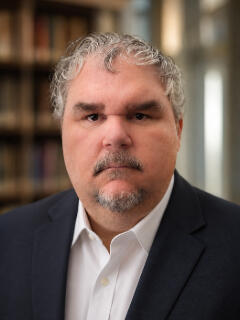 Professional academic headshot of Matt Pierce wearing a dark blazer and white shirt, photographed against a softly blurred libra Matt Pierce in a dark blazer and white shirt, photographed against a softly blurred academic background.