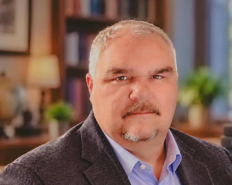 Professional academic headshot of Matt Pierce wearing a dark blazer and white shirt, photographed against a softly blurred libra Matt Pierce in a dark blazer and white shirt, photographed against a softly blurred academic background.